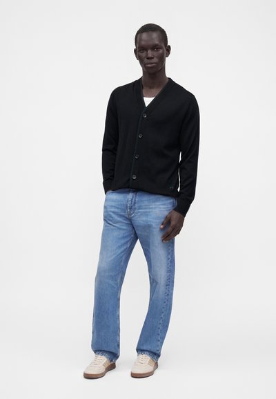 Homme debout portant un cardigan noir boutonné, un jean bleu clair, une chemise blanche en dessous et des baskets blanches sur un fond blanc uni.