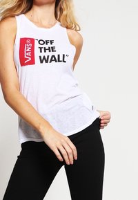 Bílý top bez rukávů s černým textem "VANS 'OFF THE WALL'" na přední straně. Měkká, lehká látka s volným střihem a zakulaceným lemem.