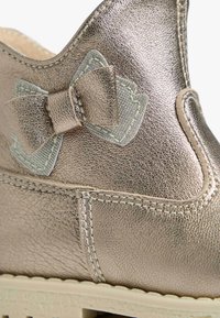 Metallisch bronzefarbenen Schuh mit strukturiertem Finish, versehen mit einer dekorativen grauen Schleife und kontrastierenden Nähten entlang der Kante und der Sohle.