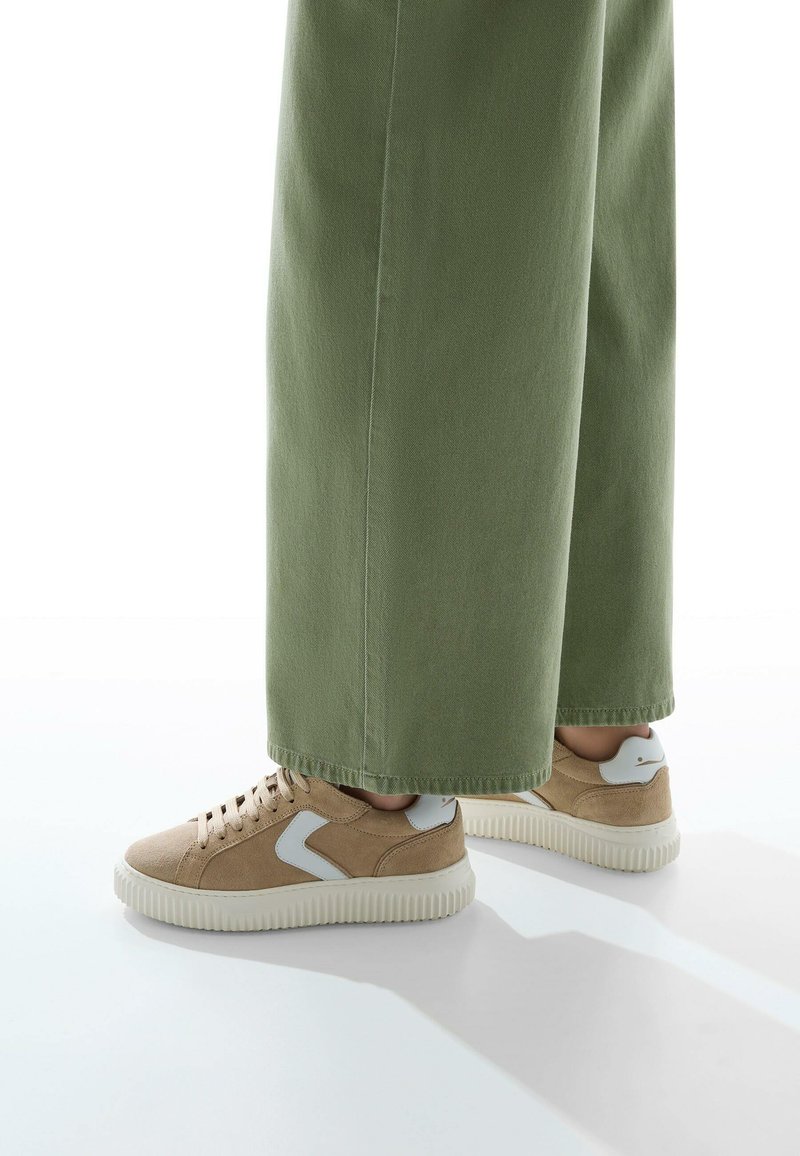 Sneaker in suede beige con dettagli bianchi, suola testurizzata e design low-top, abbinate a pantaloni larghi verde oliva.
