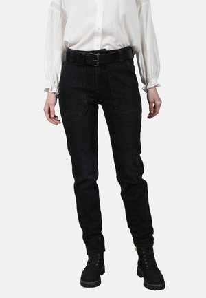 Jeans en denim noir avec une coupe slim, taille haute, grandes poches avant et une ceinture noire. Associés à un blouse blanche à manches longues et des bottes noires.