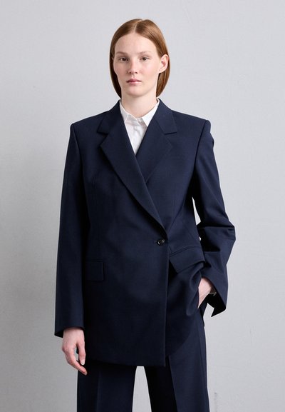 Marineblå skræddersyet blazer med store revers, enkelt knaplukning og forlommer. Båret over en hvid skjorte med krave.