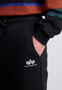 Sorte sweatpants med lommer og snor. Har et hvidt "Alpha Industries" logo og er accentueret af en sweatshirt med brune, blå og grønne striber.