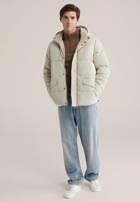 Joven vestido con una chaqueta de plumas beige sobre un suéter marrón con cremallera, jeans azul claros y zapatillas blancas, de pie con las manos en los bolsillos.