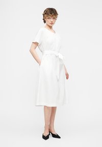 Γυναίκα με κοντά σγουρά μαλλιά, φοράει λευκό φόρεμα μέχρι το γόνατο με ζώνη στη μέση και μαύρες μυτερές γόβες, στέκεται μπροστά σε λευκό φόντο.