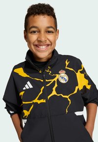 Zwarte jas met ritssluiting en gele bliksempatronen, met een Adidas-logo en Real Madrid-embleem op de borst. Elastische boorden en zakken.