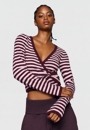 Jeune femme portant un pull cache-cœur rayé violet et rose avec un détail anneau, posant les bras croisés devant un fond uni.