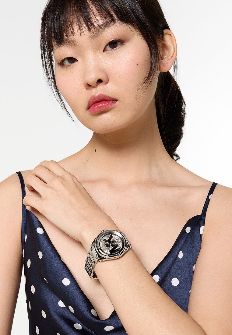 Zilverkleurig horloge met een ronde wijzerplaat, voorzien van een gegoten dial en een metalen schakelarmband. Model draagt een navy satijnen jurk met stippen.