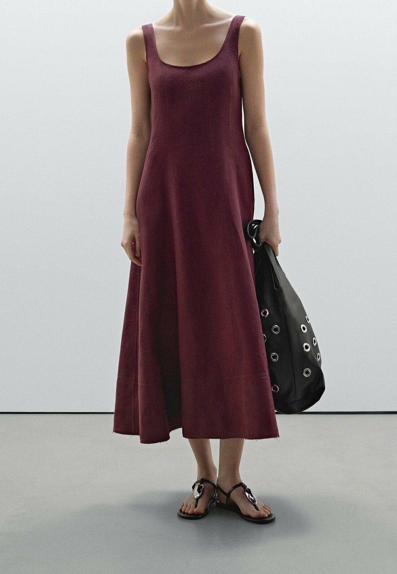 Femme portant une robe midi bordeaux sans manches, des sandales noires et tenant un grand sac noir avec des œillets argentés.