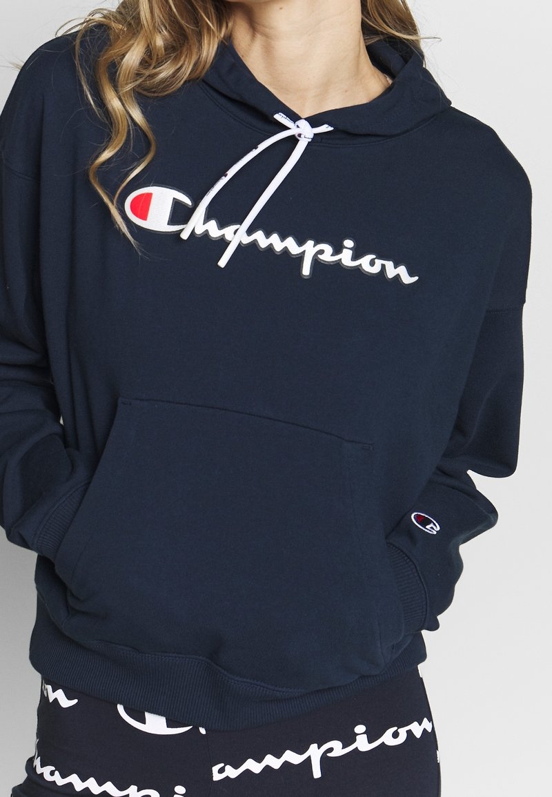 Sweatshirt à capuche bleu marine en tissu doux, avec une poche avant, des cordons blancs, et un logo "Champion" blanc proéminent avec un accent rouge.
