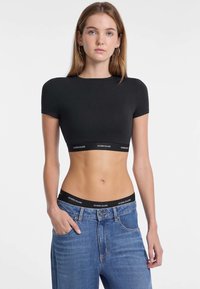 T-shirt nera corta con maniche corte, realizzata in materiale morbido. Presenta un orlo elastico brandizzato nero. Abbinata a jeans blu a vita alta.