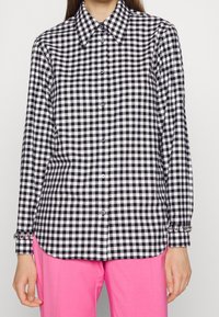 Chemise à carreaux vichy noir et blanc à manches longues, fermeture par boutons et détails de poignets ornés. Associée à un pantalon rose.