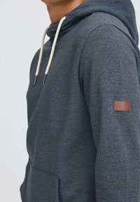 Felpa con cappuccio grigio scuro, dotata di tasca a marsupio, coulisse regolabile in beige chiaro e un piccolo patch in pelle marrone sulla manica.