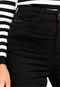 Svarta jeans med en slät yta, som har en subtil glans, framfickor med små nitar och en skräddarsydd passform.