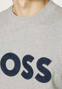 Sudadera gris con cuello redondo que presenta grandes letras bordadas de estilo vaquero en azul oscuro que dicen "OSS". Textura de tela suave.