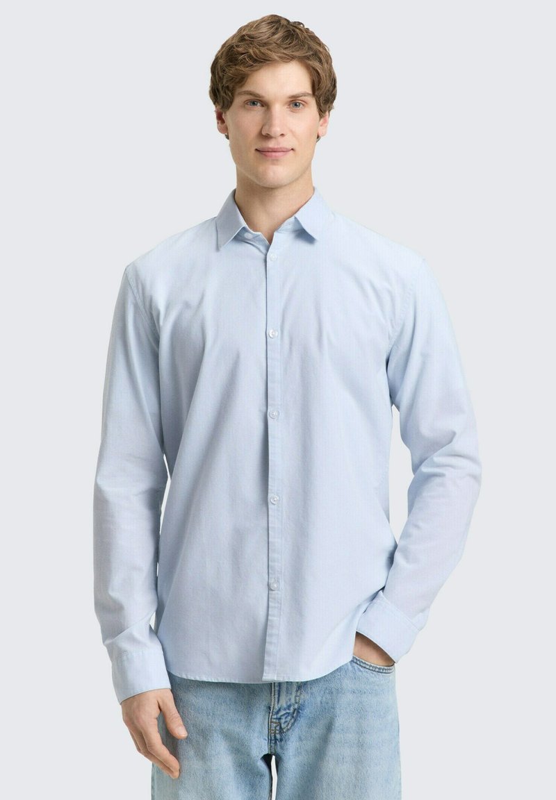 Jongeman met krullend haar die een lichtblauw overhemd met knopen en lichtblauwe jeans draagt, staand met één hand in de zak.