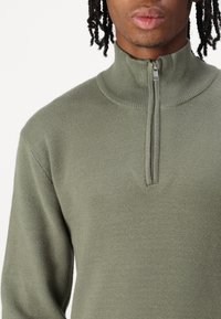 Olivgrüner Zip-Kragen-Pullover aus glattem Strickstoff, mit hohem Kragen und schlanker Passform, versehen mit einem sichtbaren Metall-Reißverschluss.