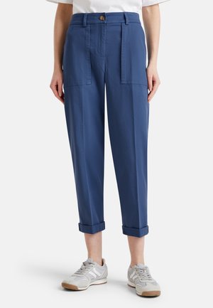 Vrouw die blauwe, hooggetailleerde, cropped broek met opgerolde boorden draagt, grote voorzakken met kleppen, een knoop, ritssluiting en grijze sneakers.