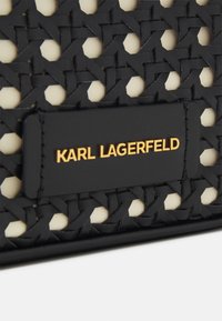 Material tejido negro con acentos crema, que presenta una etiqueta rectangular negra grabada con "KARL LAGERFELD" en letras doradas.