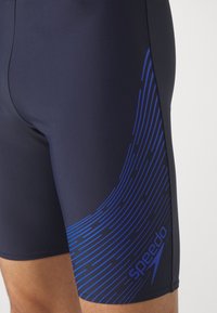 Navy svømmedrakter laget av elastisk materiale med et blått linjemønster og en Speedo-logo på den nedre delen av benet. Tett passform med glatt tekstur.