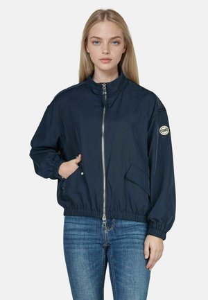 Young woman wearing a navy zip-up jacket with elastic cuffs and hem, one hand in pocket, paired with blue jeans against a plain background.