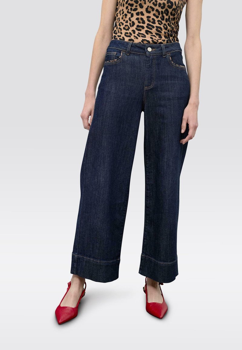 Leveälahkeiset tummat farkut korkealla vyötäröllä, joissa näkyvät saumat, etutaskut ja haalistunut lahkeen reuna. Yhdistetty punaisiin teräväkärkisiin kenkiin.