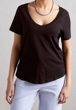 Kobieta ubrana w czarny t-shirt z krótkim rękawem i dekoltem w serek, srebrny skręcany naszyjnik łańcuszkowy, jasnoniebieskie spodnie i srebrne bransoletki na prawej ręce.