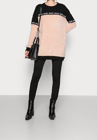 Übergroßer, flauschiger Pullover in Pfirsich und Schwarz mit Bündchen, kombiniert mit schwarzen, eng anliegenden Hosen und Stiefeletten mit Absatz. Hält eine schwarze Handtasche.