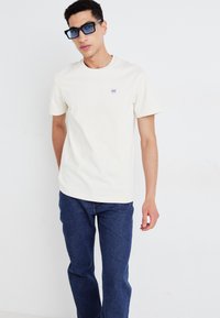 Nuori mies, joka on pukeutunut vaaleanbeigeen lyhythihaiseen t-paitaan, jossa on pieni rintalogot, tummansiniset farkut ja suuret mustat nelikulmaiset aurinkolasit, seisoo valkoista seinää vasten.