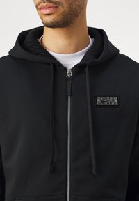 Melna zip-up kapuce jaku no mīkstas auduma, ar strukturētu, teksturētu kapuci un izteiktu logo uzkrāsu uz krūšu.