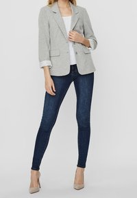Blazer grigio con texture e maniche arrotolate, due tasche frontali, indossato sopra una camicia bianca e abbinato a jeans blu scuro e tacchi beige.