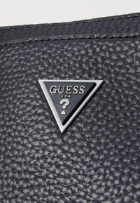 Svart texturerad läder närbild med en triangelformad metallisk GUESS-logotyp med ett frågetecken, som framhäver varumärkesidentitet och design.