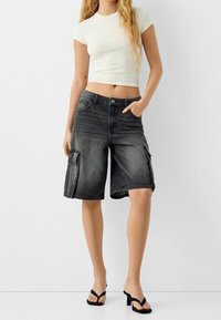 Femme portant un short cargo en denim noir délavé, un t-shirt blanc court et des sandales noires à talons, debout devant un fond blanc.