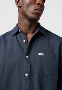 Bărbat purtând o cămașă Wrangler bleumarin cu nasturi albi, guler vizibil și un buzunar la piept cu etichetă de marcă.