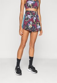 Nainen, jolla on päällään perhoskuosiset marjanpunaiset urheilushortsit ja siihen sopiva toppi, mustat Adidas-sukat ja mustat Adidas-juoksukengät.