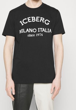 Personne portant un t-shirt noir avec un texte blanc "ICEBERG MILANO ITALIA since 1974" et un pantalon bicolore avec jambes noires et blanches.