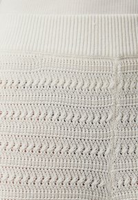 Krémová pletenina s texturovaným, otevřeným cik-cak vzorem a žebrovaným pasem. Design je lehký a vzdušný.