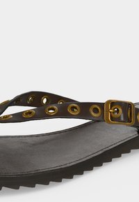 Σανδάλι σκούρου καφέ με ρυθμιζόμενο λουρί που διαθέτει χρυσές μεταλλικές τρύπες και αγκράφα, εξωτερική σόλα με ανάγλυφη επιφάνεια για καλύτερο κράτημα.