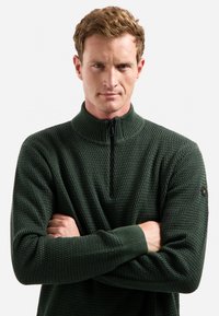 Man met kort haar in een donkergroen gestructureerd half-rits trui, met gekruiste armen, tegen een eentonige lichte achtergrond.