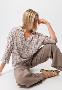 Gemusterte Bluse mit geometrischem Print in Hellbraun und Weiß, mit V-Ausschnitt und 3/4-Ärmeln, kombiniert mit lockeren sandfarbenen Hosen und Rutsch-Sandalen.