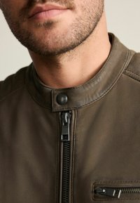 Braune Lederjacke mit hohem Kragen, schwarzem Reißverschluss und einer Reißverschlusstasche auf der linken Seite. Glatte Textur, minimalistisches Design.