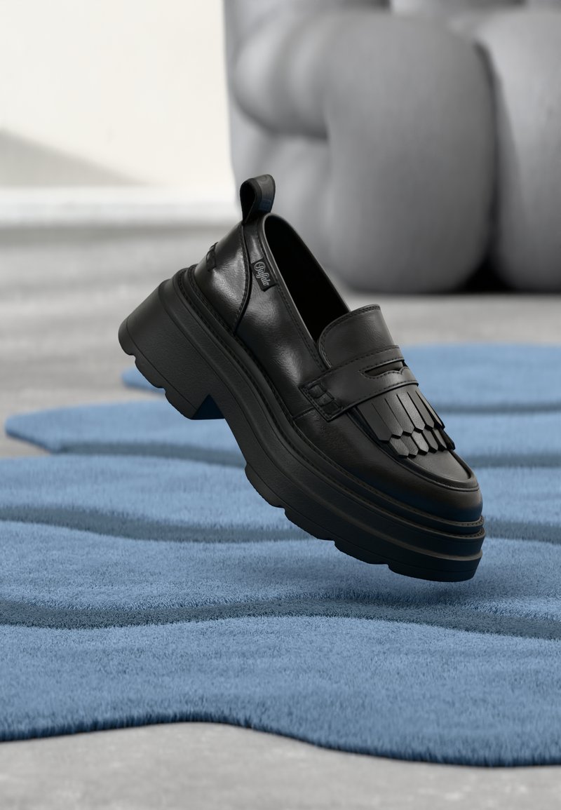 Svart skinn slip-on sko med frynseaccent, tykk såle og teksturert finish, plassert på et blått teksturert teppe.