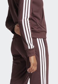 Brun atletisk treningsoverall med hvite tre-stripe detaljer. Har en slim fit, ribbede mansjetter og en glidelåslomme på toppen.