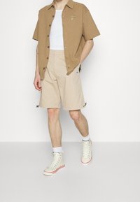Chemise courte à manches courtes marron clair avec des boutons sur le devant, shorts beiges avec des ajusteurs latéraux, et baskets montantes blanches avec des accents rouges.