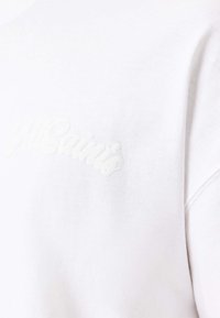 Tissu blanc avec une broderie subtile en relief portant l'inscription "MSaints" en écriture cursive sur la partie supérieure de la poitrine.