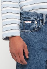 Acercamiento de una mano al lado de unos vaqueros de mezclilla azul con una etiqueta de Calvin Klein, que lleva una camisa de manga larga con rayas en azul claro, blanco y gris.
