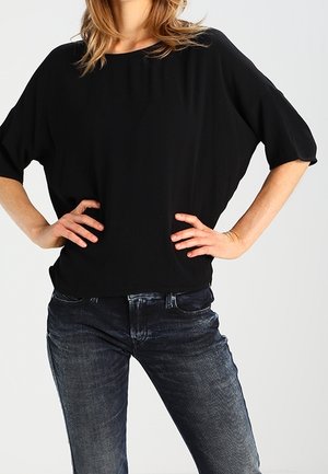 Vrouw draagt een loszittend zwart kort mouwen topje en verbleekte zwarte jeans met de handen rustend op de heupen tegen een effen achtergrond.