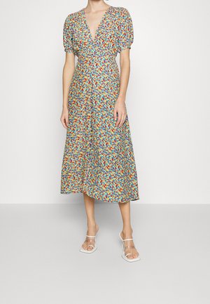 Femme portant une robe fleurie mi-longue à manches ballon et décolleté en V profond, associée à des sandales à talons hauts à brides transparentes, debout contre un mur blanc.