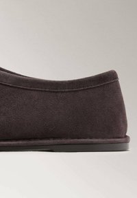 Zapato de ante gris oscuro con un diseño minimalista y un talón redondeado y elegante. La suela es plana y tiene una textura sutil. No hay herrajes ni acentos visibles.