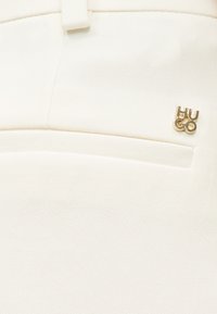 Κοντινό πλάνο λευκού υφάσματος με τσέπη σχισμής και μικρό χρυσό μεταλλικό λογότυπο με την επιγραφή "HUGO" στη δεξιά πάνω πλευρά.